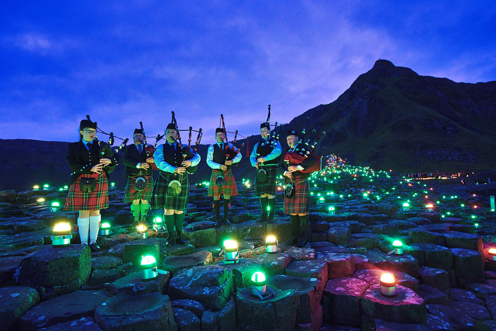 Six Northern Irish bagpipers play amongst low lights on the ground at Gian's Causeway